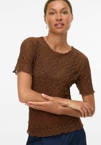 Kvinna klädd i en brun texturerad kortärmad topp med vågiga kanter och mörkblå byxor, poserar med armsluten på vit bakgrund.