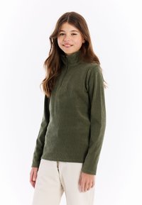 Meisje met lange bruine haren, gekleed in een olijfgroene, geribbelde sweatshirt met rits en lichtbeige broek, glimlachend tegen een witte achtergrond.
