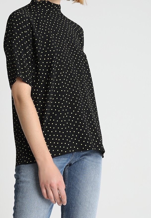 Svart kortärmad topp med hög halsringning, prydd med ett gult prickmönster och en avslappnad passform. Bärs tillsammans med ljusblå denimjeans.