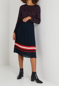 Top navy a maniche lunghe con piccolo motivo floreale rosso, abbinato a una gonna plissè navy con righe rosse e bianche, e stivali neri alla caviglia.