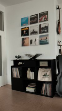 Svart treskap med vinylplater, en dekorativ vase og en kremfarget veske. På veggen vises albumcover, inkludert "Oasis."