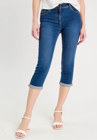 Pantalon capri en denim bleu foncé avec une coupe ajustée, ourlets retroussés et taille mi-haute, associé à des sandales blanches à talons blocs.