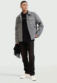 Mann in grauer gesteppten Jacke, schwarzen Hosen, schwarzem Shirt und weißen Sneakers, der einen schwarzen Fransen-Schal hält und zur Seite schaut.