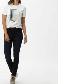 Vit kortärmad t-shirt med abstrakt grön och grå grafisk tryck, i kombination med mörka skinny jeans och beige sneakers.