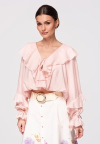 Femme portant un chemisier à volants rose clair à manches longues et une jupe blanche à imprimé floral avec une ceinture beige à grande boucle circulaire.