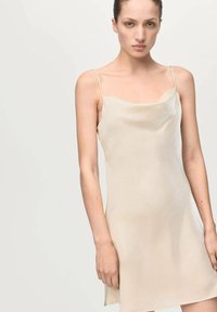 Vestido lencero en beige claro, con finas tiras y una silueta suavemente drapeada. Textura suave con un brillo sutil, corte hasta la rodilla.