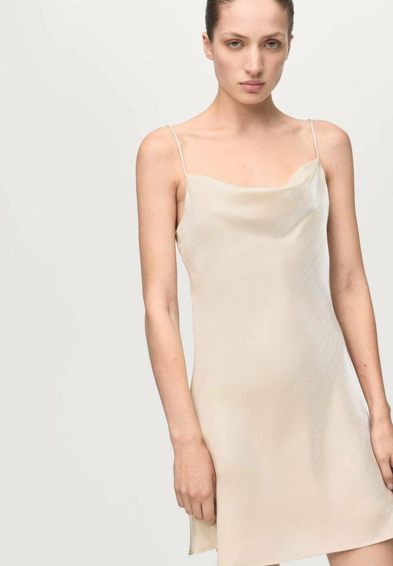 Vestido lencero en beige claro, con finas tiras y una silueta suavemente drapeada. Textura suave con un brillo sutil, corte hasta la rodilla.