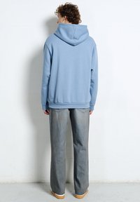 Osoba o kręconych włosach, ubrana w jasnoniebieską bluzę z kapturem, szare dżinsy i beżowe buty, stojąca twarzą do gładkiej białej ściany.