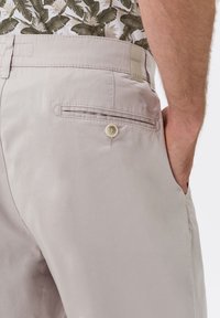 Muž v světlobéžových kalhotách s knoflíkovou zadní kapsou a košili s květinovým vzorem, ruka v boční kapse.