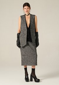 Gilet sans manches texturé gris sur un haut noir à boutons, assorti d'une jupe, de gants noirs et de chaussures à plateforme.