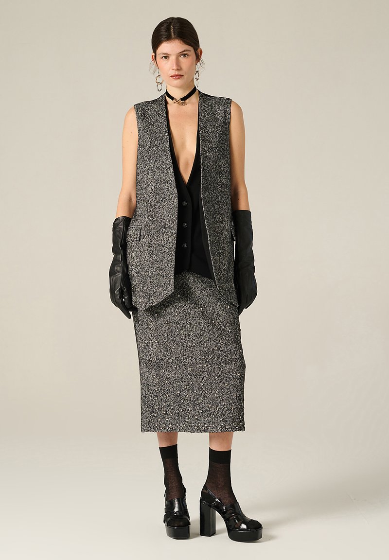 Gilet sans manches texturé gris sur un haut noir à boutons, assorti d'une jupe, de gants noirs et de chaussures à plateforme.