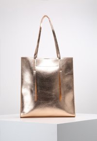 Metaliczna torba tote w kolorze różowego złota, z mocnymi płaskimi uchwytami, gładką teksturą, prostokątnym kształtem oraz bez widocznych zapięć lub ozdób.