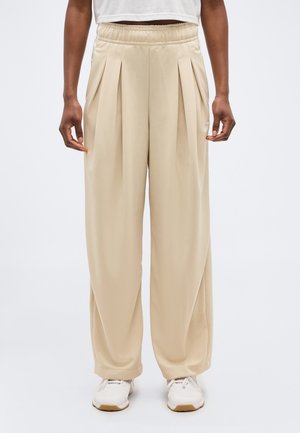 Inimene kannab beeže volditud laiapõlvelisi pükse elastse vöökohaga ja valgeid tossusid, taust on lihtne.