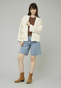 Veste style kimono beige clair avec un motif texturé, portée sur un top marron et un short en denim bleu. Des bottines tan complètent la tenue.