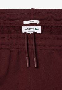Vinröda tapered fit sweatpants med elastisk midja, dragsko och Lacoste-logotag. Slät textur, avslappnad design, unisex stil.