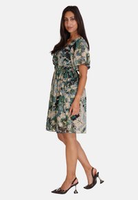 Robe fleurie dans des tonalités de vert, crème et bleu, avec une taille cintrée, des manches courtes et une longueur arrivant au genou. Portée avec des escarpins noirs à brides.