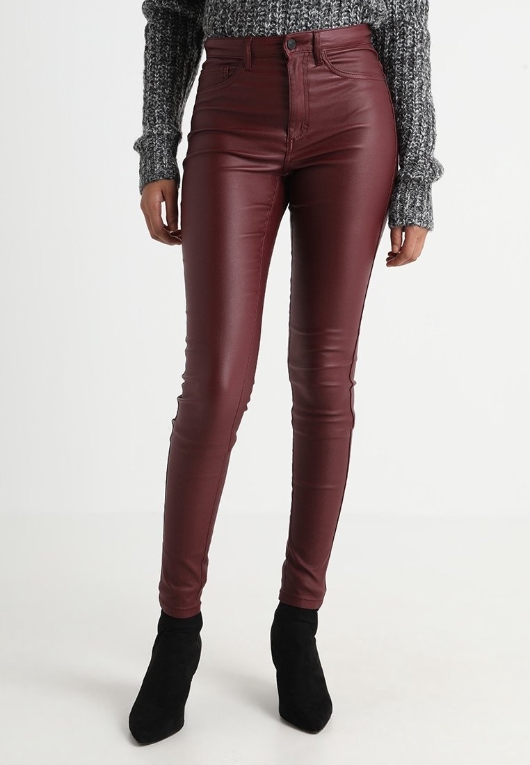 Femme portant un pantalon en cuir bordeaux moulant et taille haute, associé à un pull en maille gris et des bottines noires sur un fond uni.
