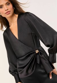 Kobieta w czarnej satynowej bluzce na zakładkę z bufiastymi długimi rękawami i metaliczną klamrą w talii.