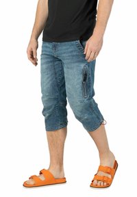 Denim-Capri-Hosen in einem verblassten Blauton, mit seitlicher Reißverschlusstasche und Kordelzug am Saum, kombiniert mit orangen Slidesandalen.