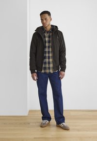 Modell trägt eine schwarze Kapuzenjacke, ein Karohemd in Blau und Beige, blaue Jeans und graue Sneakers mit weißen Streifen. Hintergrund aus Holzboden.