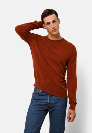 Brown crew-neck sweater made of soft fabric with long sleeves and a slightly asymmetrical hem, paired with blue jeans.
