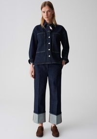Giacca in denim blu scuro con grandi tasche e chiusura a bottoni, abbinata a pantaloni coordinati con orli in denim più chiaro.