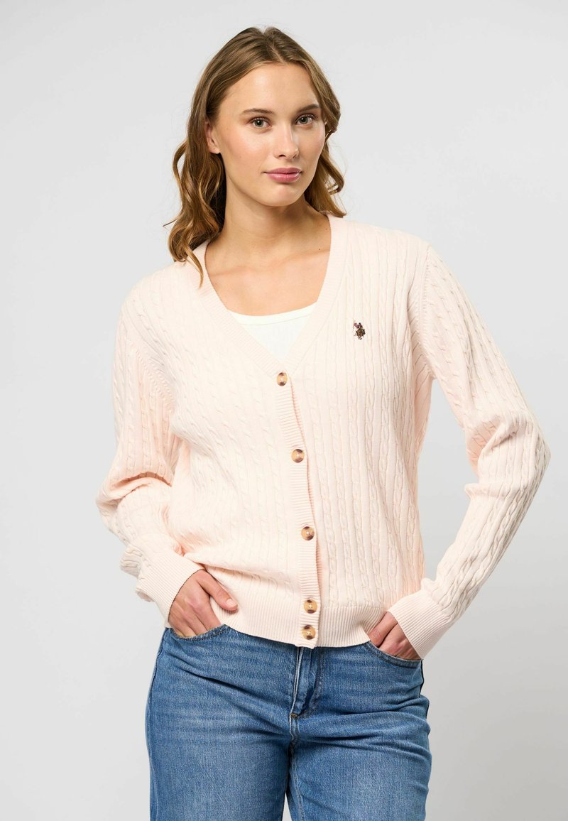 Cardigan en tricot rose clair avec un motif en câble, décolleté en V, cinq boutons sur le devant, poignets côtelés et un petit badge brodé sur la poitrine.