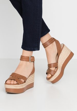 Sandales marron à talon compensé avec des sangles cloutées, des détails en jute et une plateforme épaisse. Elles disposent d'une sangle au niveau de la cheville avec une boucle en argent.