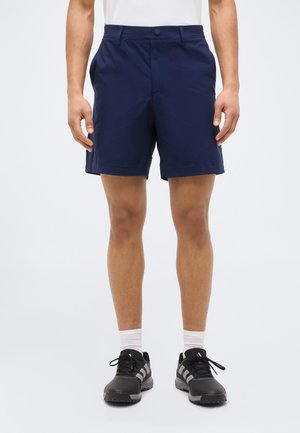 Uomo che indossa pantaloncini blu navy, calzini bianchi e scarpe da ginnastica nere, in piedi con le braccia rilassate ai lati contro uno sfondo uniforme.