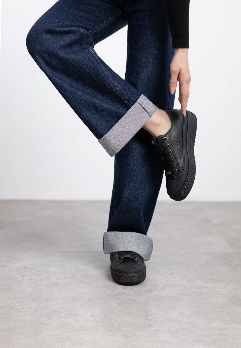Zapatillas negras con punta redonda, diseño de cordones y suelas texturizadas, combinadas con vaqueros azul oscuro remangados en el dobladillo y forro interior gris.