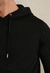 Hanorac negru cu glugă și șnur, având o textură tricotată moale și accente metalice de culoare argintie la capetele șnurului.