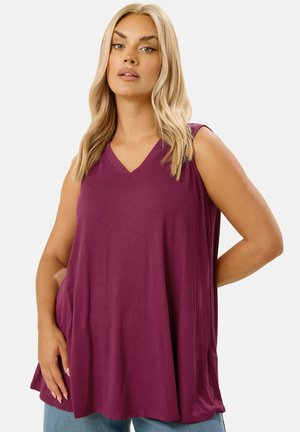 Femme blonde portant un haut sans manches ample côtelé de couleur prune et un jean bleu, debout avec une main dans la poche.