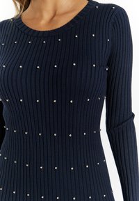 Marineblaues, geripptes Kleid mit langen Ärmeln, ausgestattet mit gleichmäßig verteilten metallischen Nieten auf dem Stoff. Rundhalsausschnitt und eine taillierte Silhouette.