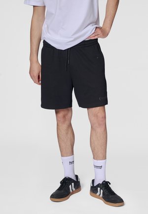 Schwarze Shorts mit elastischem Bund und Kordelzug, kombiniert mit weißen Socken und schwarzen Sneakers. Die Shorts sind dezent gebrandet.