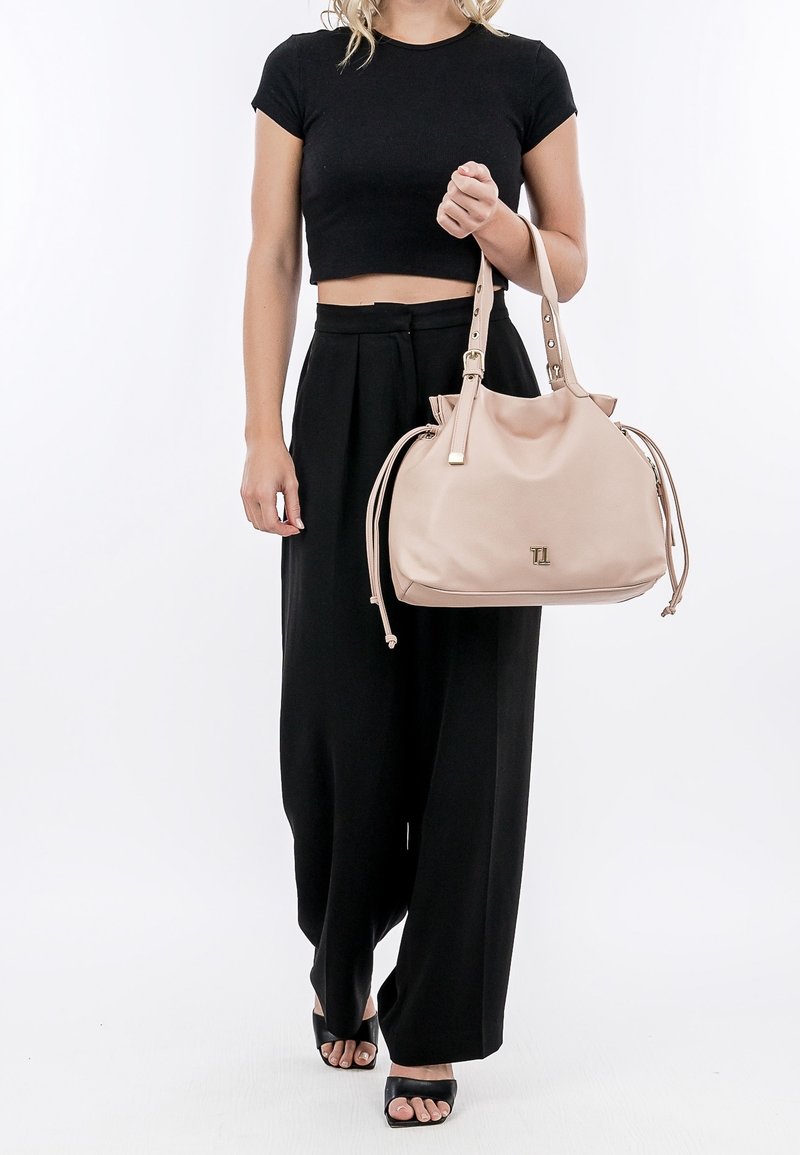 Pink leather handbag with gold hardware, drawstring sides, and a logo on the front, held by a person wearing a black crop top and wide-leg pants.