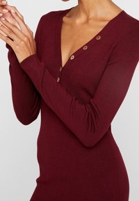 Robe en maille côtelée bordeaux avec un décolleté en V profond, des manches longues et trois boutons au niveau de l'encolure. Texture lisse, silhouette ajustée.