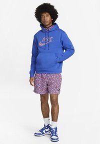 Blå hætte sweatshirt med et grafisk logo, parret med røde og blå mønstrede shorts. Iført med hvide sokker og blå sneakers.