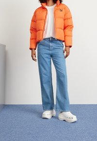 Giubbotto piumino arancione, t-shirt bianca, jeans blu a vita alta e zoccoli bianchi spessi. L'ambientazione presenta un pavimento blu con una parete neutra.