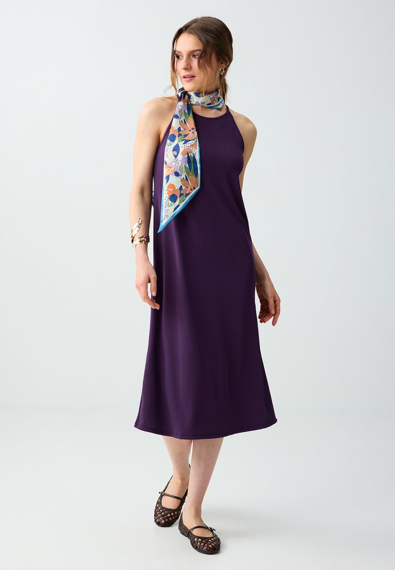Femme en robe midi sans manches violet foncé, portant un foulard fleuri coloré autour du cou et des ballerines noires tressées, debout devant un fond uni.