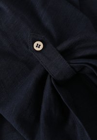 Tela de lino azul marino con una textura suave, que presenta un botón de madera como cierre. La camisa está ligeramente fruncida cerca de la zona del botón.