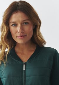 Green padded jacket with a ribbed round neck and zipper closure, featuring a textured surface and smooth fabric.