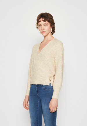 Beige innpakningsgenser laget av mykt, teksturert materiale med V-hals og sidebånddetalj, kombinert med blå jeans.