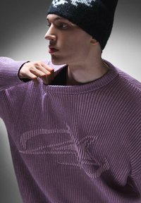 Maglione viola a costine con una vestibilità morbida, caratterizzato da un motivo testurizzato sul davanti. Il modello indossa un berretto di maglia nero.