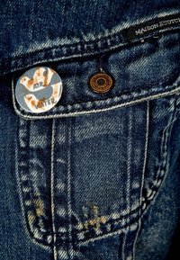 Denim-tasku, jossa on messinkirivetti ja kuvioitu nappi, jossa lukee "ILMA" ja "VESI". Kankaan väri on haalistunut sininen ja siinä on lievää kulumista.