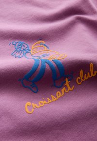 Lila Stoff mit einer gestickten Cartoonfigur, die ein Croissant hält. Darunter steht in Gelb "Croissant-Club".