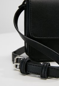 Czarna skórzana torba na ramię o teksturowanym wykończeniu, z srebrną klamrą i regulowanym paskiem. Kompaktowy, prostokątny kształt.