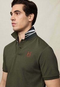 Muž v olivově zeleném polo tričku se proužkovaným límečkem vyhrnutým nahoru a červeným logem "H Hackett" na hrudi, dívající se vlevo na jednotném pozadí.