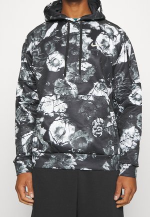Mand iført en sort og hvid Nike hættetrøje med blomstertryk, forlomme og sorte snore, kombineret med sorte bukser.