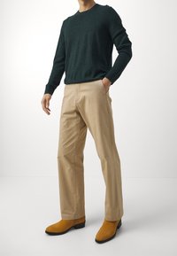 Person som bär beige raka byxor, senapsfärgade mockakängor och en mörkgrön långärmad tröja, står med en hand i fickan.