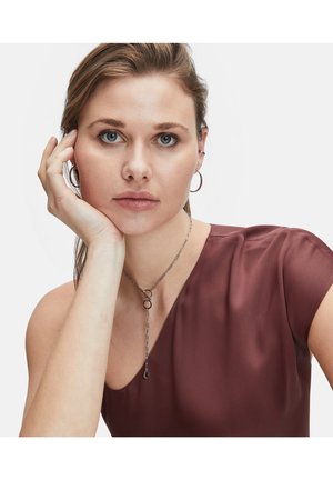 Silberne Kettenhalskette mit kreisförmigem Akzent, getragen mit einem kastanienbraunen Seidentop mit asymmetrischem Ausschnitt. Strukturiertes Haar, neutrales Makeup.